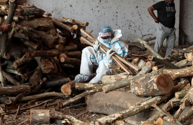 A family member wearing personal protective equipment (PPE) carries wood to prepare a funeral pyre for their relative at a cremation ground