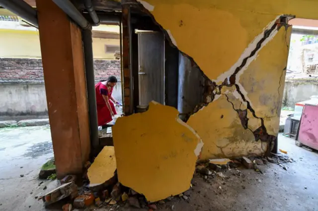 A woman collects water from a tap near a collapsed wall