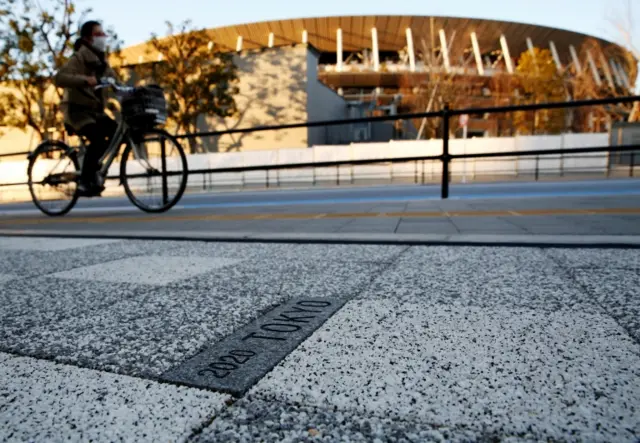 A woman cycles past a sign for Tokyo 2020 Olympic Games on the pavement in front of the National Stadium, the main stadium of Tokyo 2020 Olympics and Paralympics, in Tokyo, Japan, on 3 February 2021