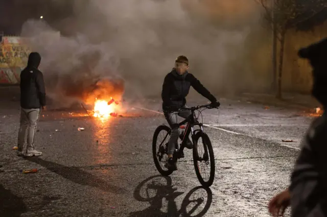 Young man on a bike with a fire behind him and a shield over his face