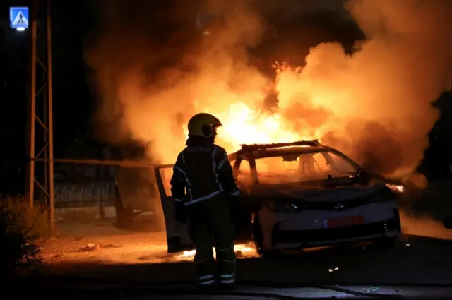 An Israeli firefighter stands near a burning police car during riots in Lod, Israel (12 May 2021)