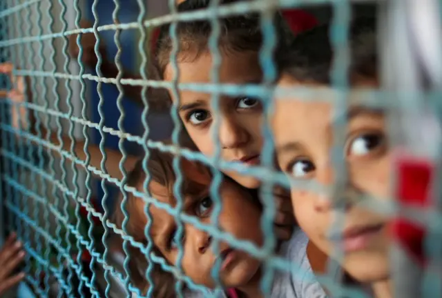 Palestinian children, who fled their homes due to Israeli air and artillery strikes, look through a window fence at a UN-run school where they take refuge, in Gaza City