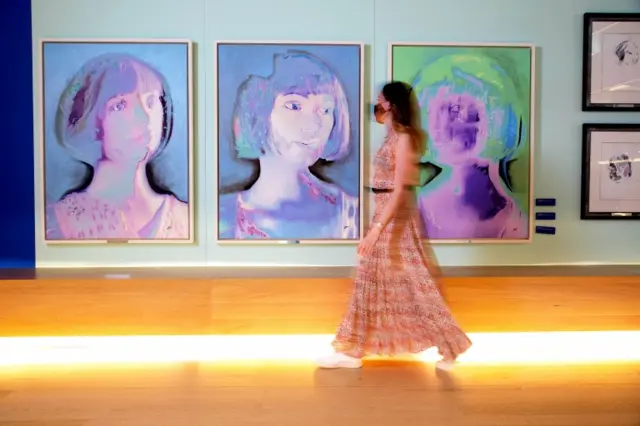 Woman walks past pictures in exhibition Ai:Da