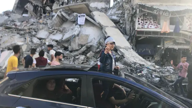 A boy wearing a keffiyeh gestures as he drives past a wrecked building in Gaza City (21 May 2021)