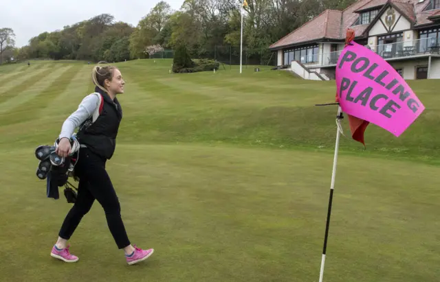 A woman walks across a golf course. A flag in one of the holes reads 'polling place'