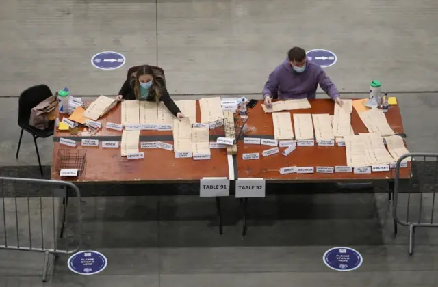 Two people count votes on a big table, with ballots placed in piles next to names of corresponding parties