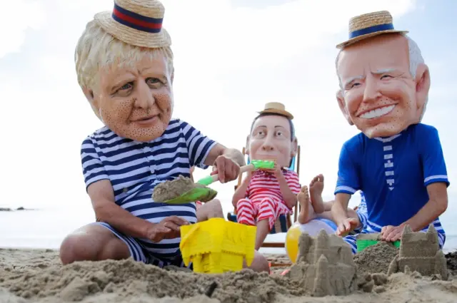 Oxfam activists wearing papier mache heads to represent G7 leaders protest on a beach near Falmouth