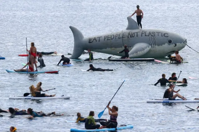 A man stands on a prop shark near the protesters on paddleboards