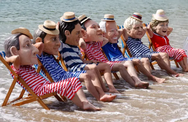 Oxfam activists wearing papier mache heads depicting G7 leaders perform during a climate protest at Swanpool Beach near Falmouth.