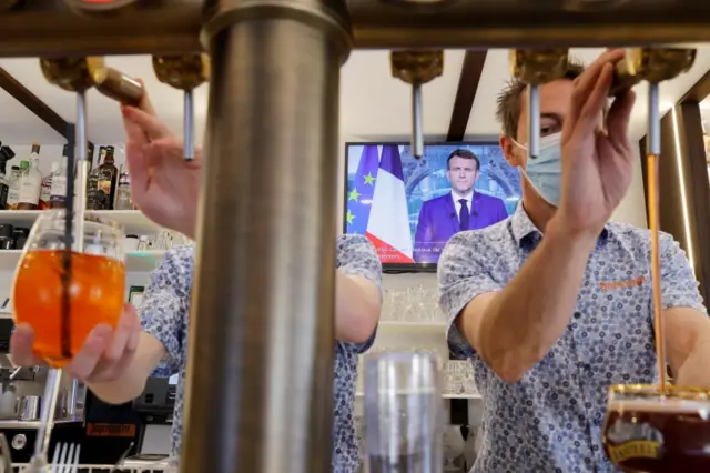 Employees pour drinks in a pub as French President Macron, on TV, addresses the nation on 12 July