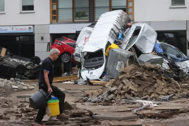 A person carrying buckets walk amid debris near a pile of damaged cars after flooding in Bad Neuenahr-Ahrweiler