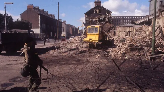 A soldier stands beside a demolished house.