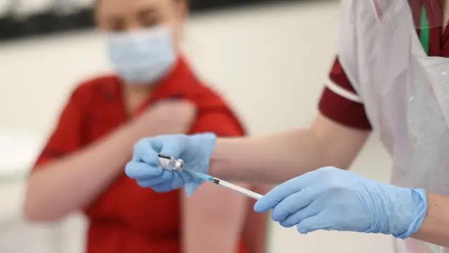 A nurse drawing the vaccine from a vial with a patient in the background with their sleeve rolled up