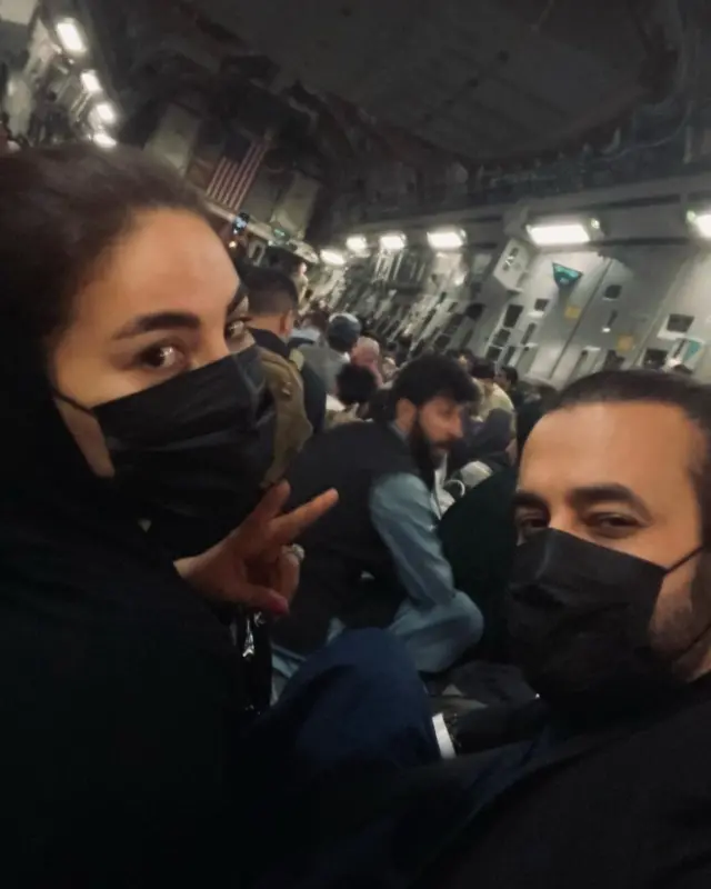 A photograph shows Sayeed and a male, wearing masks, sitting on floor of packed plane