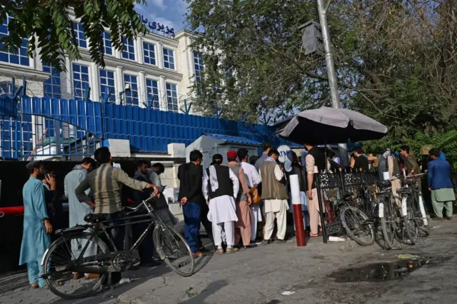 Afghan people stand in a queue as they wait for their turn to collect money from an ATM in front of a bank along a roadside in Kabul on August 21, 2021