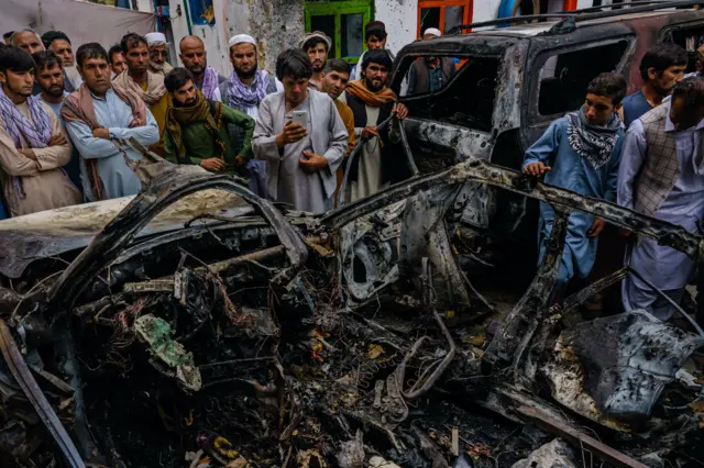 Relatives and neighbors of the Ahmadi family gathered around the incinerated husk of a vehicle targeted and hit earlier Sunday afternoon by an American drone strike, in Kabul, Afghanistan, Monday, Aug. 30, 2021