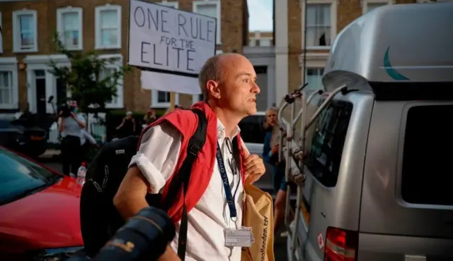 Dominic Cummings walks past cameras and a sign saying 'One rule for the elite'