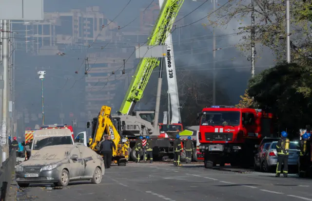 Emergency service vehicles surrounded by damaged buildings and ash covered cars.