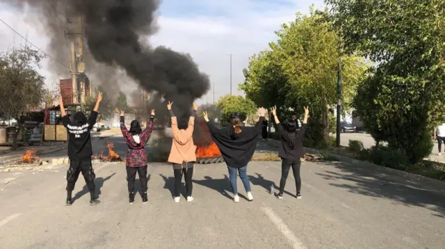 Five people standing in front of burning tyres and hold up the peace sign with their fingers