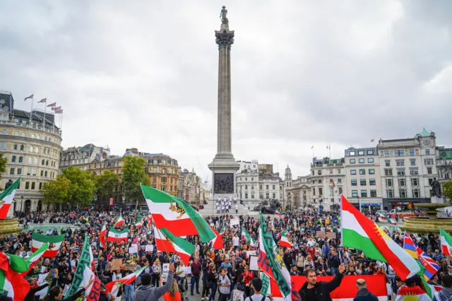 Hundreds of protestors, many with Iranian flags, standing in Trafalgar Square, London.