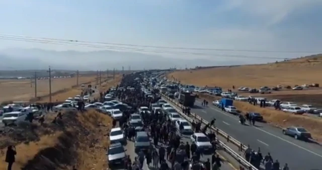 A massive traffic jam as cars line a dual carriageway. Lots of pedestrians mill about.