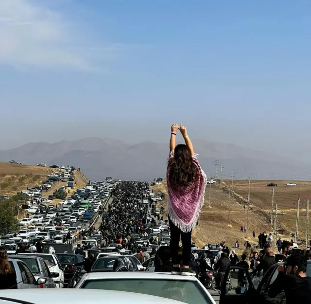 A girl wears a red shawl and stands on top of a car. In front of her is a huge traffic jam and hundreds of people stand on the road.