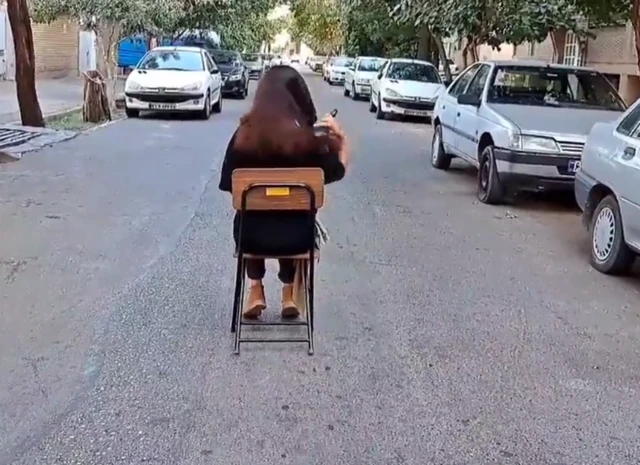 A woman sits on a chair and brushes her hair in the middle of the street in an act of defiance