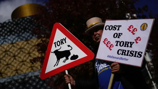 Welsh activist Steve Bray holds up anti-Conservative signs, including one that says 'Cost of Tory crisis' and one of a graphic of a bull and its excrement