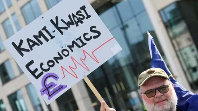 A man holds up a sign which reads "Kami-Kwasi economics"