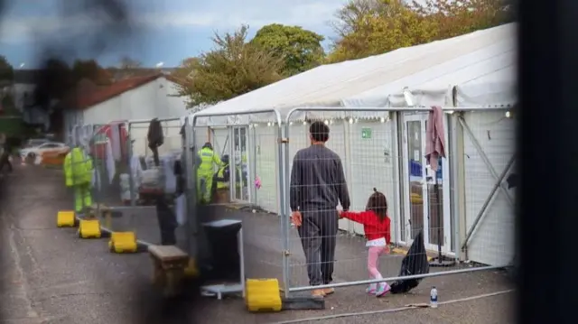 Man in outdoor holding pen at Manston holding hands with a young girl, inside metal fencing next to temporary tent structures