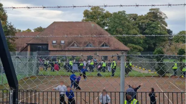 Children playing on a tennis court inside Manston, with visible barbed wire fencing in the foreground