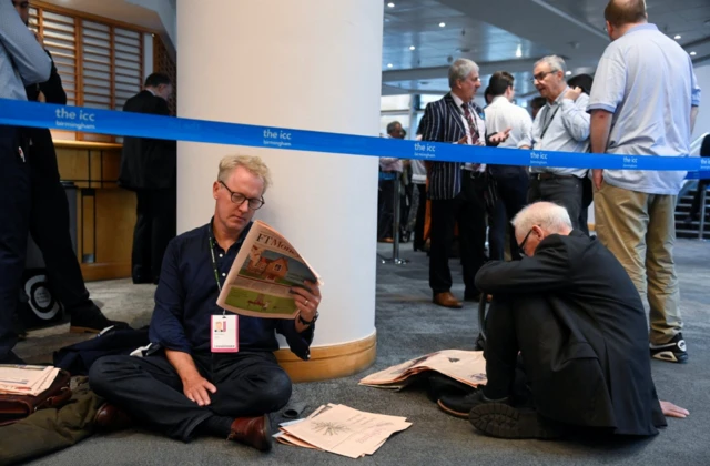 People sit on the floor as they wait in queue; one of them reads the Financial Times