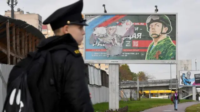 A military cadet stands in front of a billboard promoting contract army service in Saint Petersburg