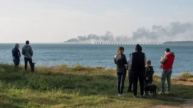 People watch fuel tanks ablaze and damaged sections of the Kerch bridge in the Kerch Strait, Crimea