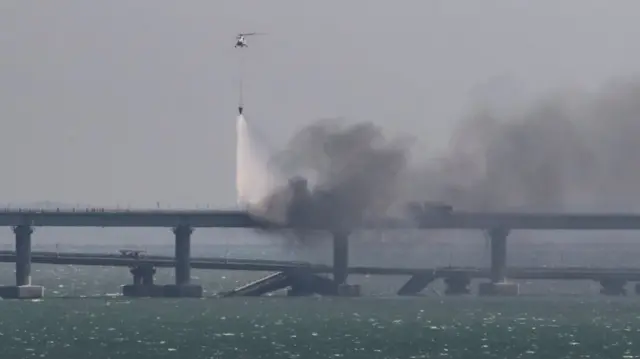 A helicopter drops water to extinguish fuel tanks on fire next to damaged sections of the bridge