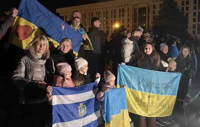 A group of Ukrainians stand with children holding Ukrainian and Kherson flags