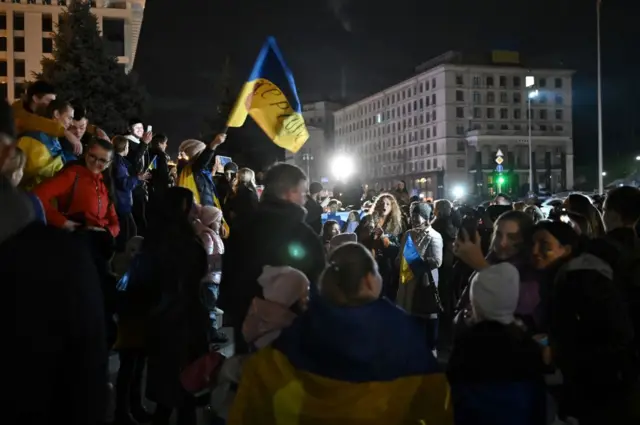 Crowds gather in the darkness waving and wearing Ukrainian flags