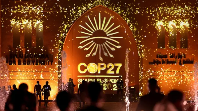 People pass in front of a wall lit with the sign of COP27 as the climate summit takes place