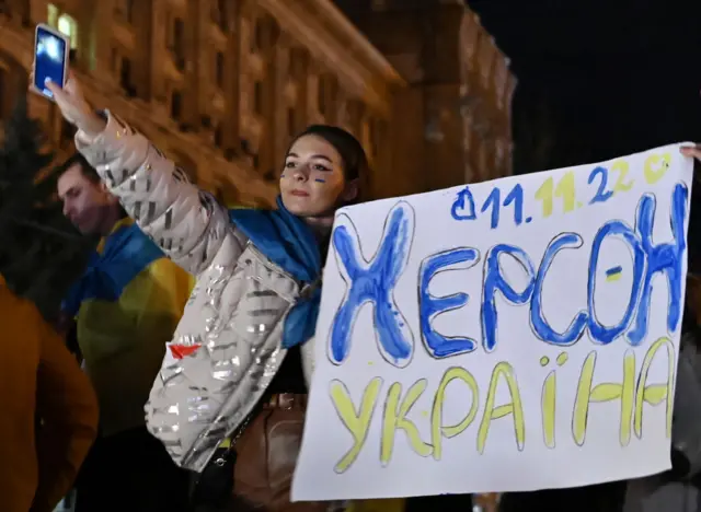 A woman draped in Ukraine's flag holds a sign which says "Kherson, Ukraine" with today's date