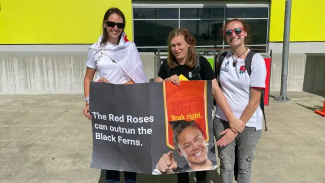 Three women, two with England shirts, and one with a sign saying the red roses can outrun the black ferns yeah right