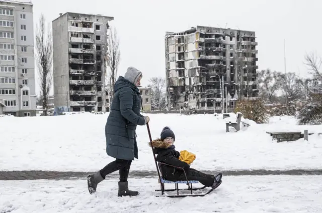 A woman pushes a child through snow in Borodyanka, in front of damaged buildings