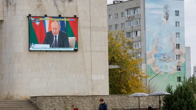 A television on the side of a building in Enerhodar, where the power plant is located, shows a broadcast of Russian President Vladimir Putin on Russian state TV. A mural on a nearby building includes a depiction of a nuclear power plant