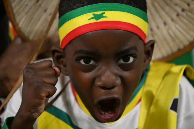 A boy with a Senegal headband, shirt and scarf cheers and clenches his fist.