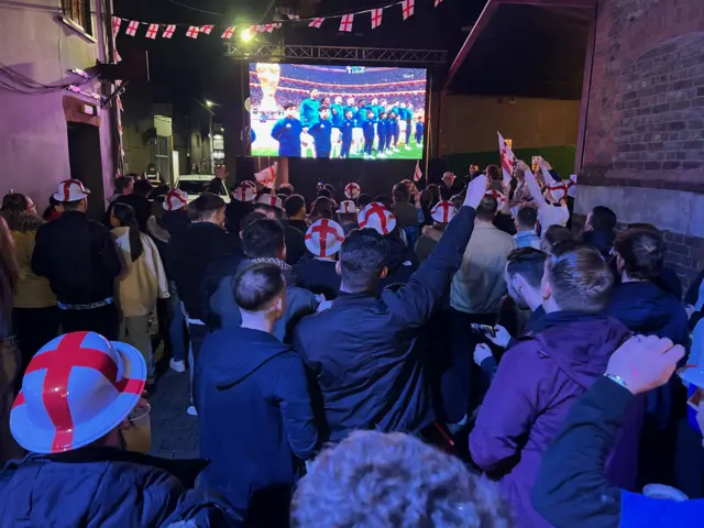 A group of people wear England hats in the street as they watch the game on the big screen