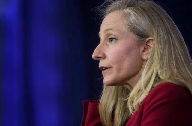 Side profile of US Representative Abigail Spanberger, who is wearing a red blazer and sat beside a blue backdrop