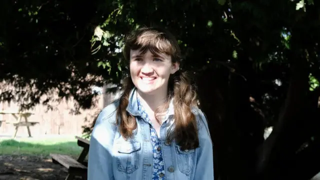 Roisin is standing in front of a tree and is wearing a denim jacket and blue dress.