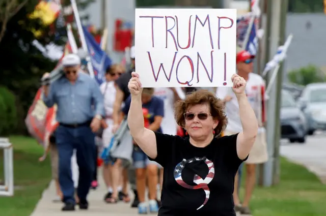 A woman in a black T-shirt with 'Q' written on it, holds up a sign saying "Trump won!"