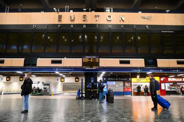 Passengers stand below a blank departure display board while they wait at the concourse of Euston station