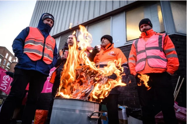 workers in high-viz jackets stand around a bin fire to keep warm