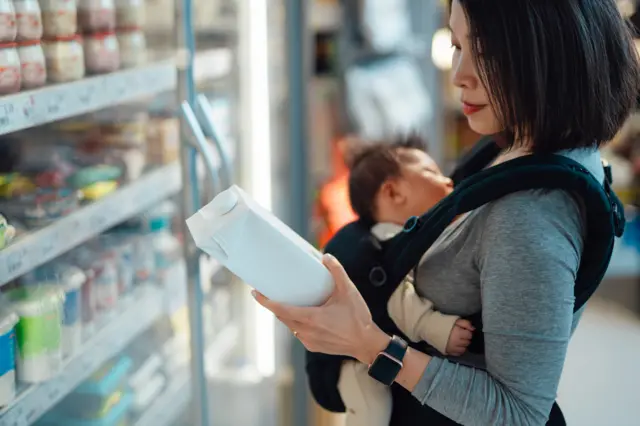 Stock image shows a woman carrying a baby in a supermarket and looking at a carton